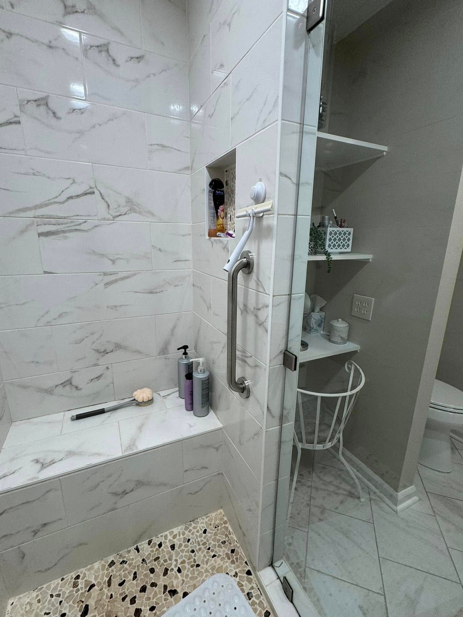 White marble shower with glass door, built-in bench, and shelves with bathroom items.