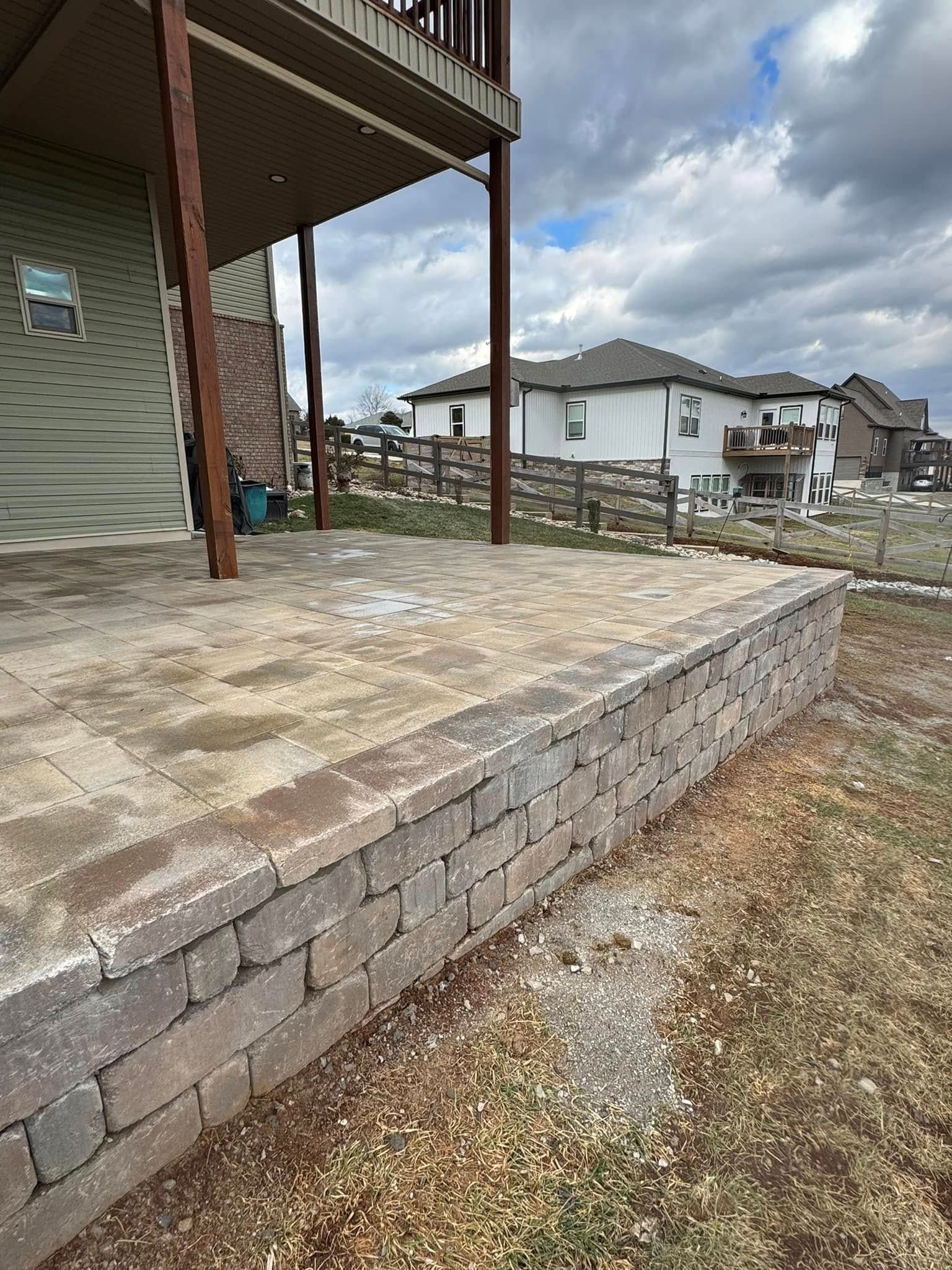 Brick patio with retaining wall, beneath a deck, in a yard. Cloudy sky, residential houses in the background.
