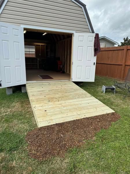 Wooden ramp leading into an open shed with white doors, on a grassy lawn.