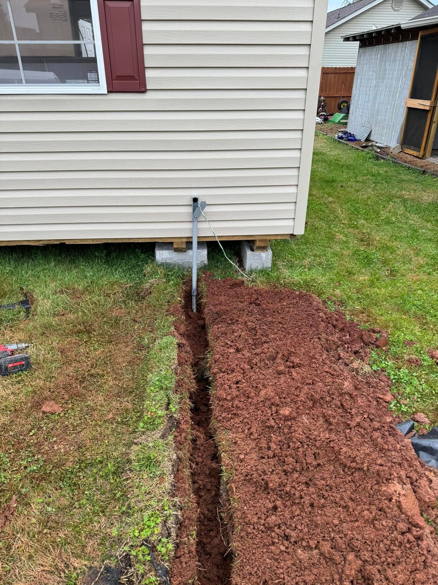 Trench dug in the yard near a beige-sided building.  Pipe protrudes from the building's wall.