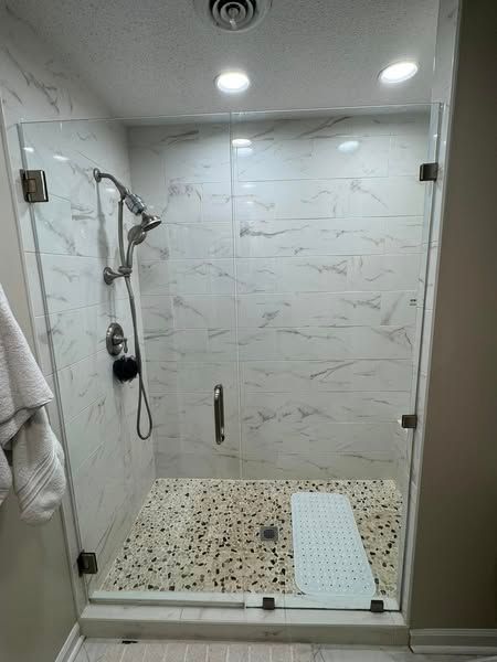Walk-in shower with marble-like tile, glass doors, and a pebble floor. A shower bench is on the right.
