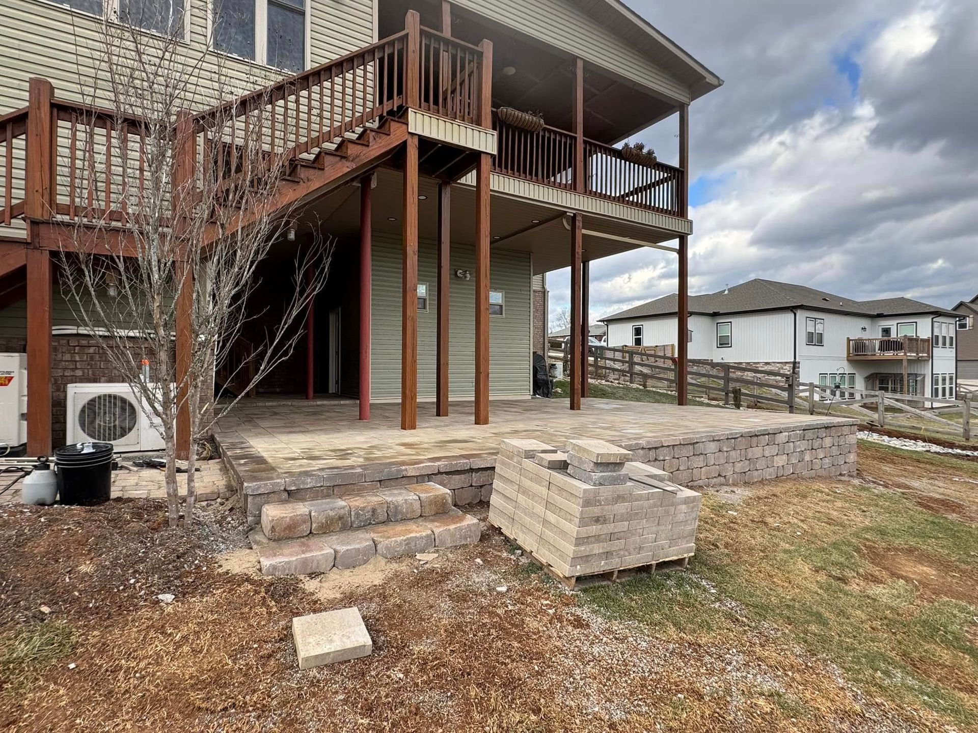 Backyard with tiered patio under a two-story deck. Brown and tan paving stones, beige siding, cloudy sky.