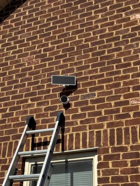A security camera and solar panel mounted on a red brick wall next to a window, with a ladder leaning against the wall.