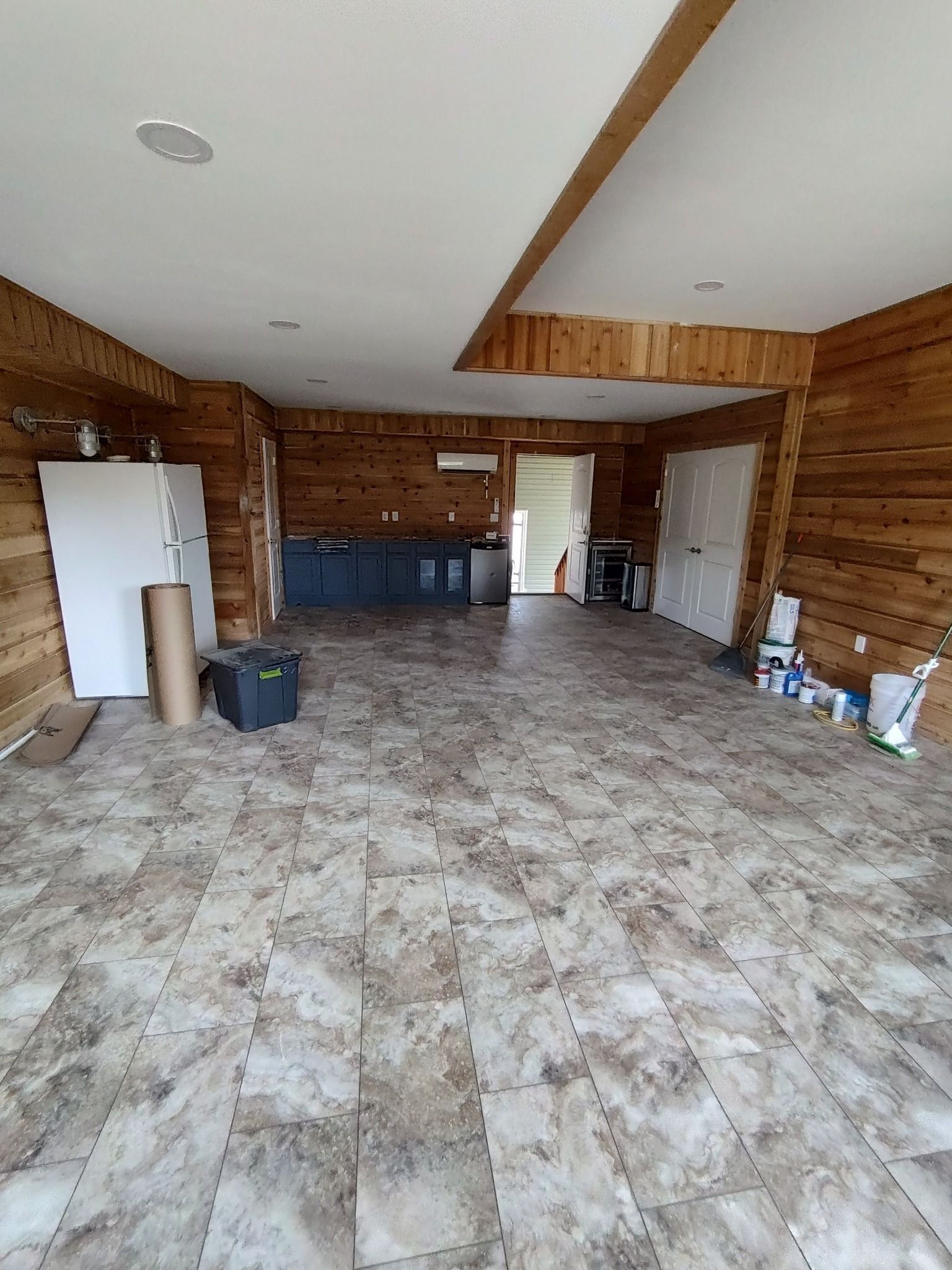 Empty room with wood paneling, tiled floor, and door. Construction materials and cabinets visible.