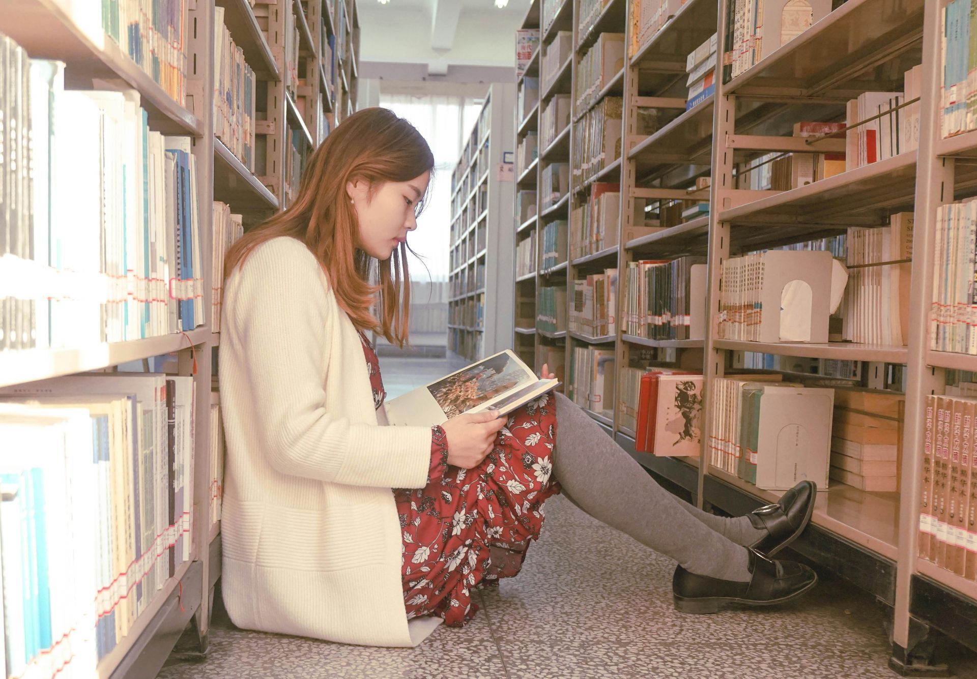Uma mulher está sentada no chão de uma biblioteca lendo um livro.