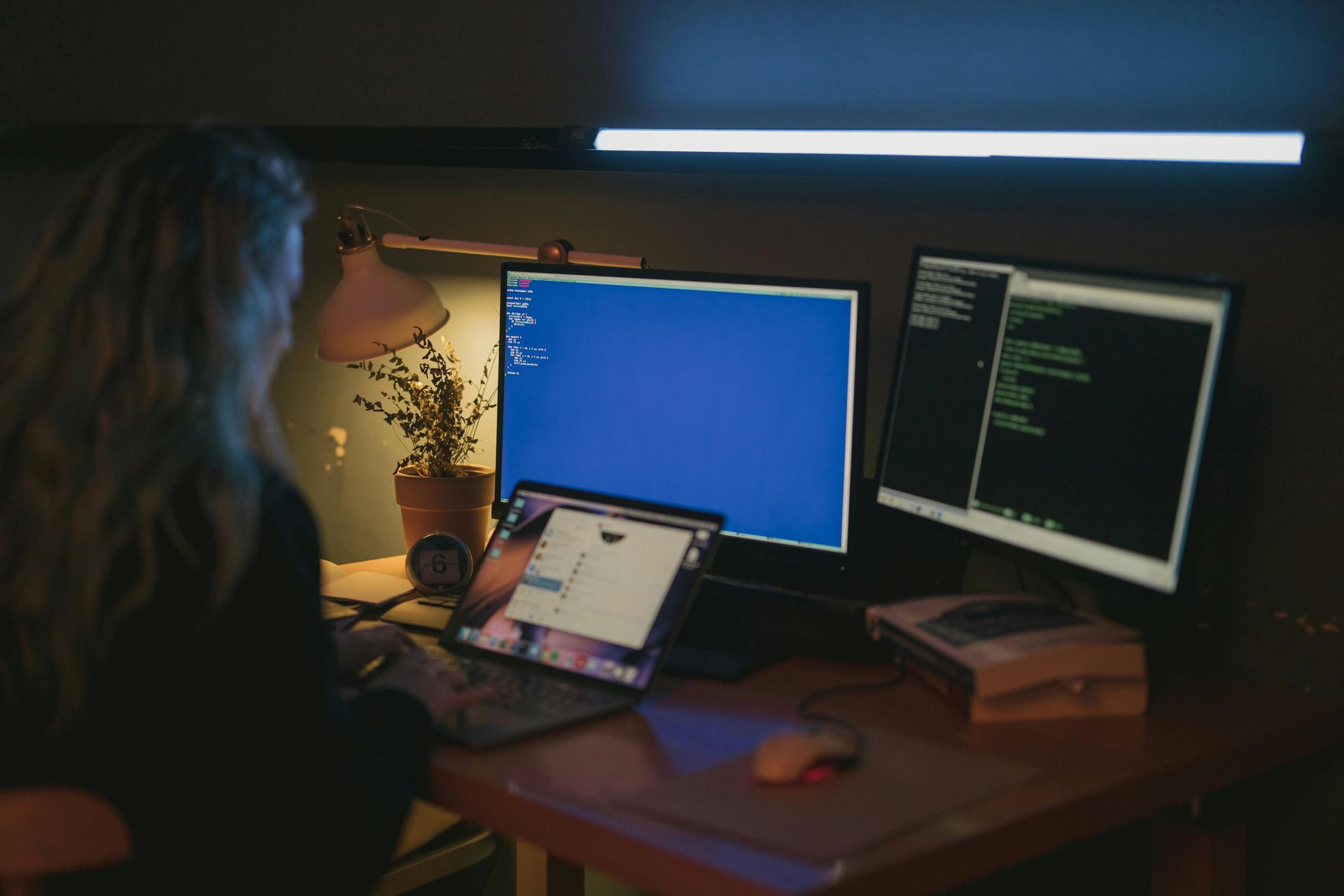 Uma mulher está sentada em uma mesa com dois monitores de computador e um laptop.