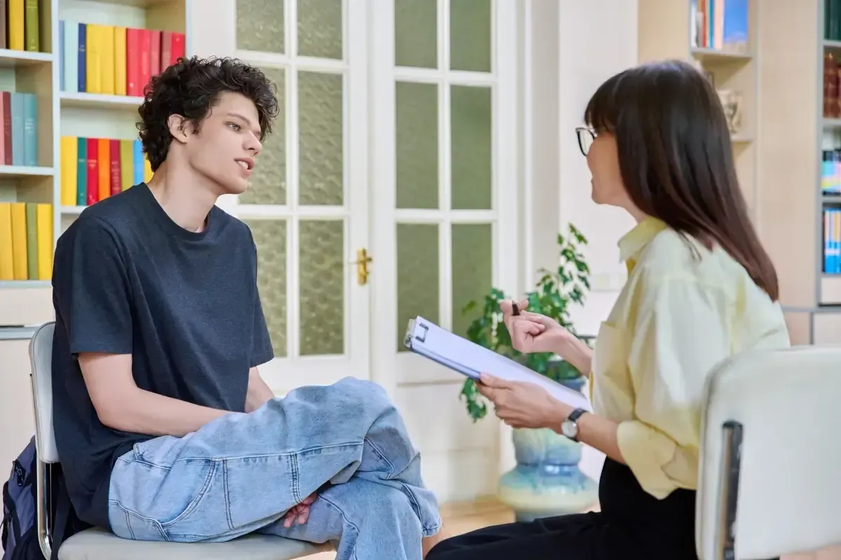 Man in therapy session talking to a woman holding a clipboard; bookshelf and a decorative door are in the background.