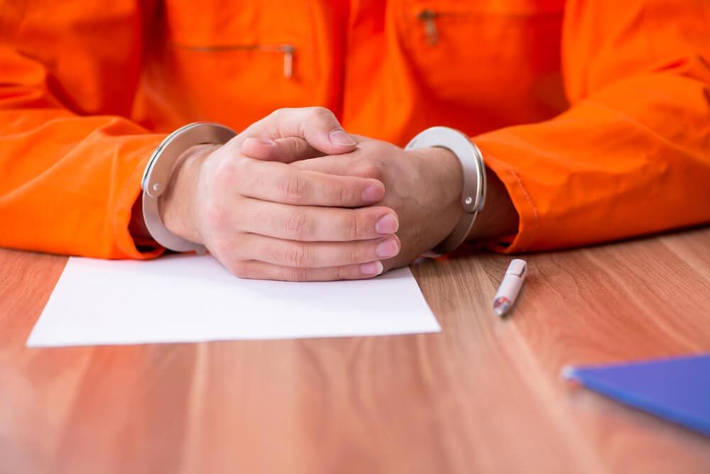 A person in an orange jumpsuit sits at a table, handcuffed, with paper and pen in front of them.
