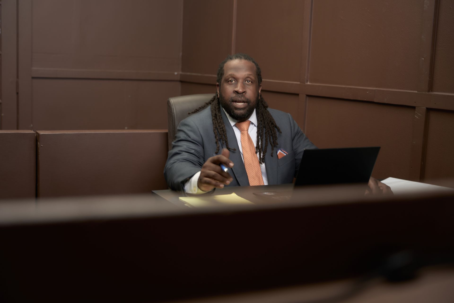 Man with dreadlocks sits at a desk in a courtroom, wearing a suit and tie, pointing.