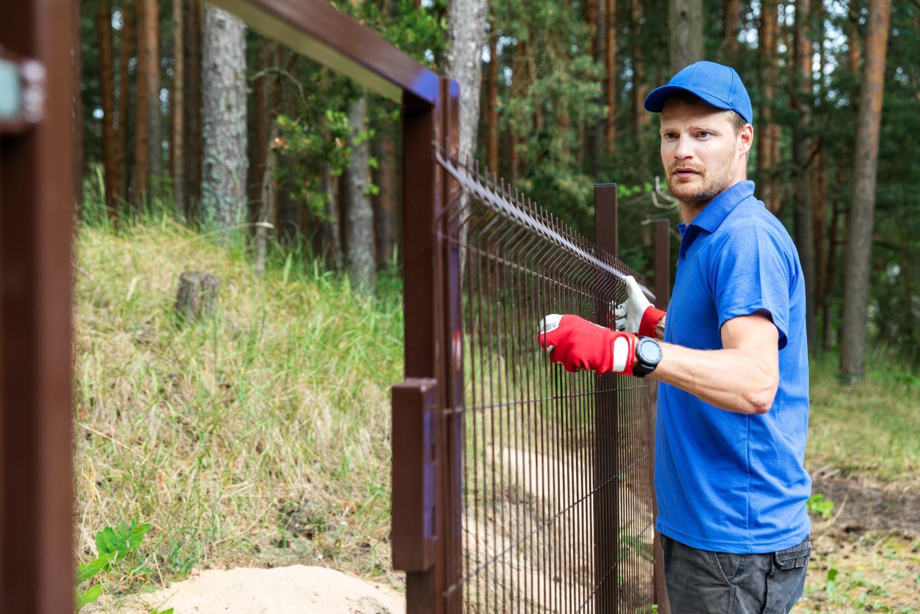 Man painting a brown metal fence outdoors, wearing blue shirt and cap, red gloves, looking at the camera.