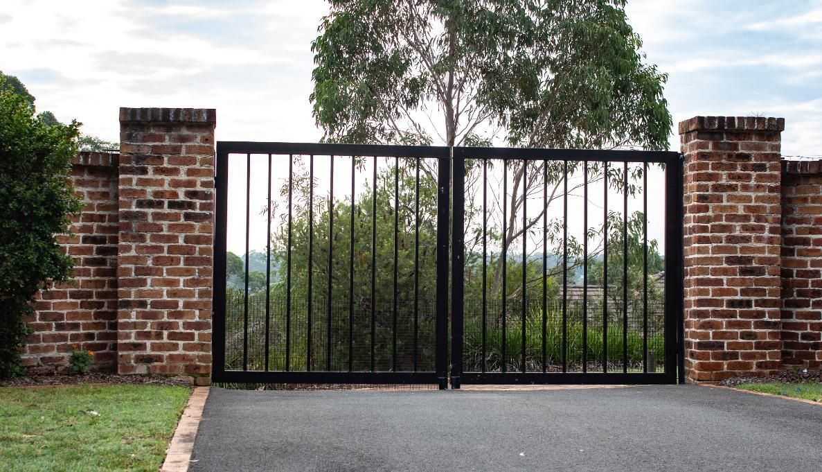 Black metal gate between brick pillars, leading to a driveway with foliage in the background.