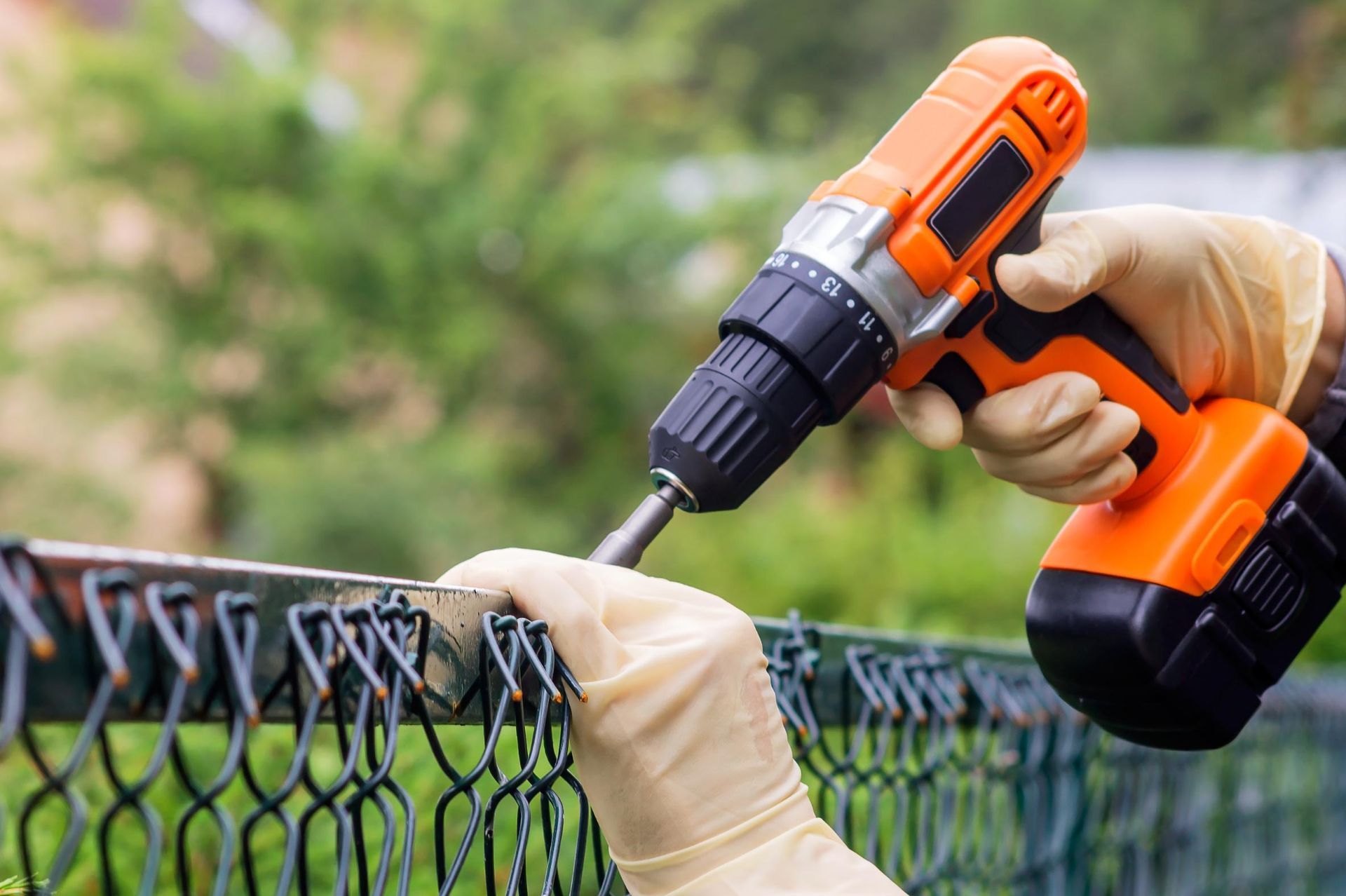 Person using a drill to secure a chain link fence, wearing gloves outdoors.