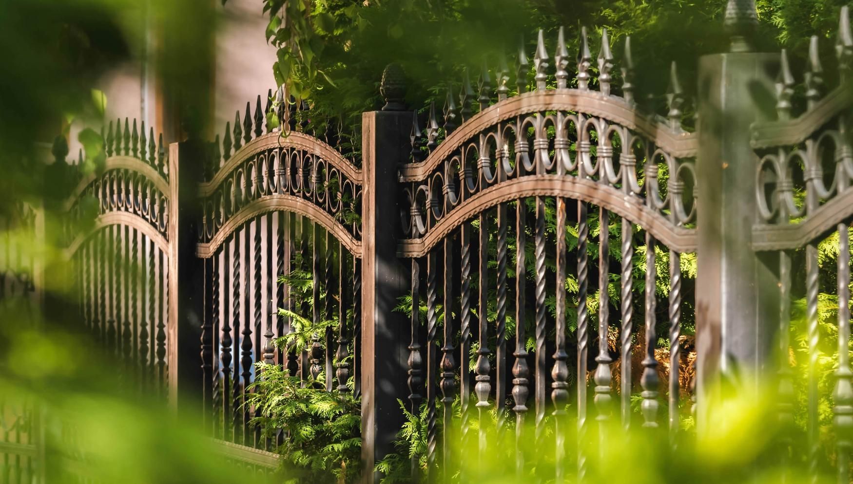 Ornate wrought-iron fence with open gates, surrounded by green foliage.