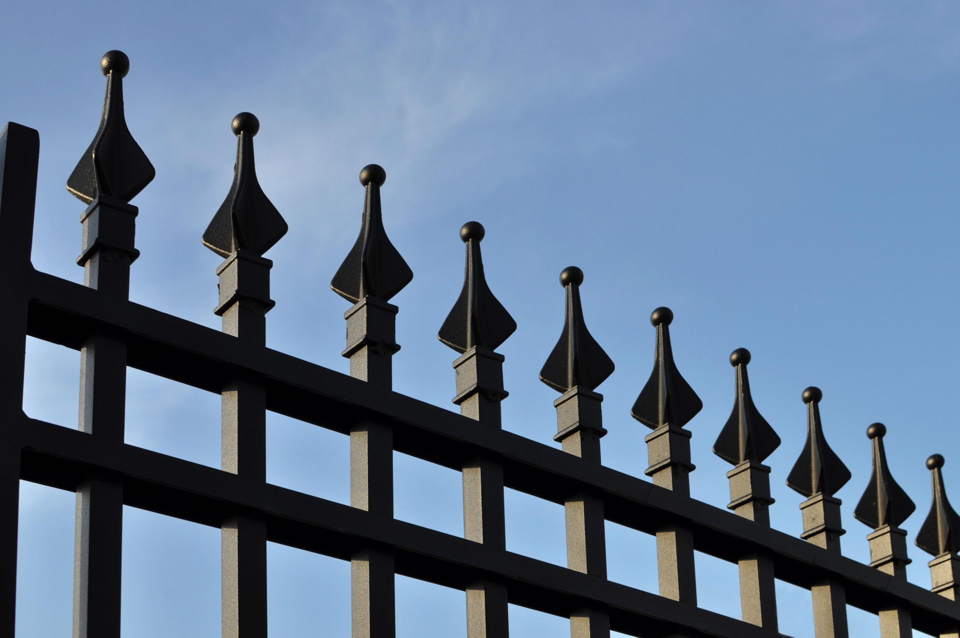 Black wrought-iron fence with decorative spear points against a blue sky.
