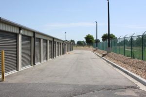 Storage units with metal doors, asphalt road, security fence, and blue sky.