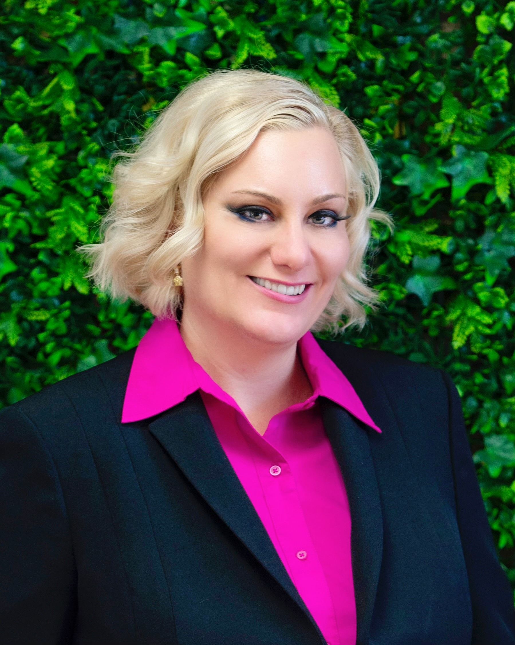 Woman in a black suit with a pink shirt smiles in front of a green ivy wall.