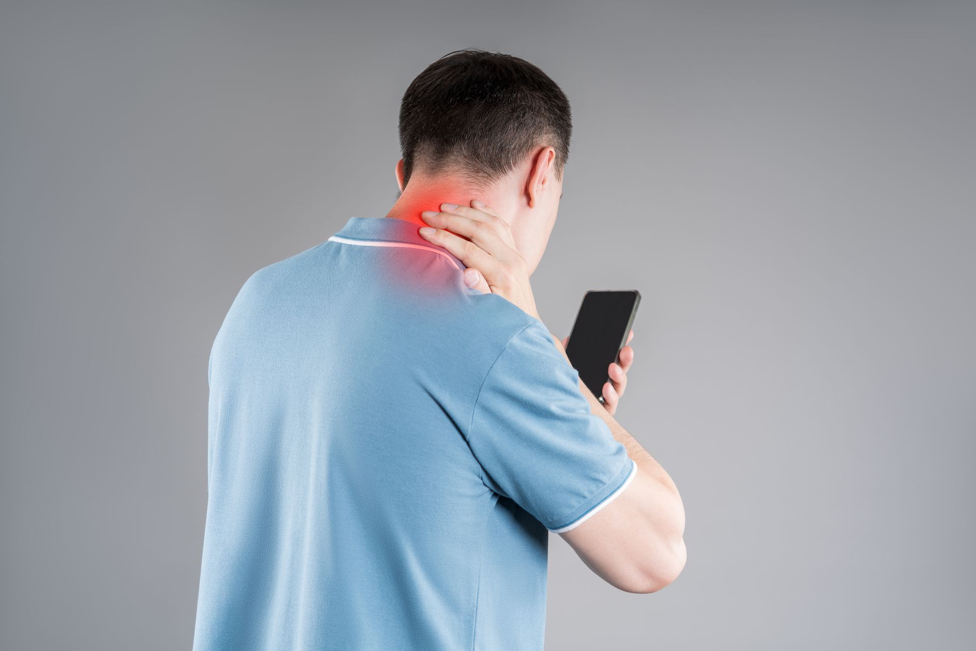 Person in a light blue shirt holding a smartphone and clutching their neck, which is highlighted in red to show pain.