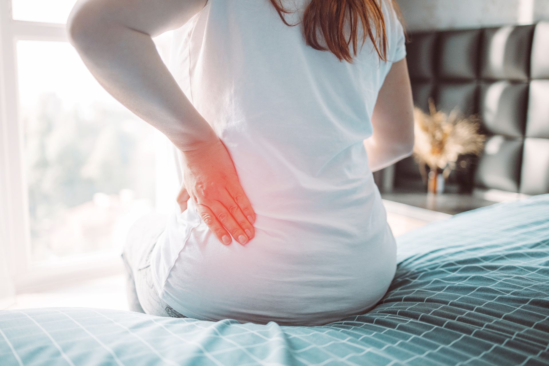 Woman sitting on bed, touching lower back in pain, with red highlight.
