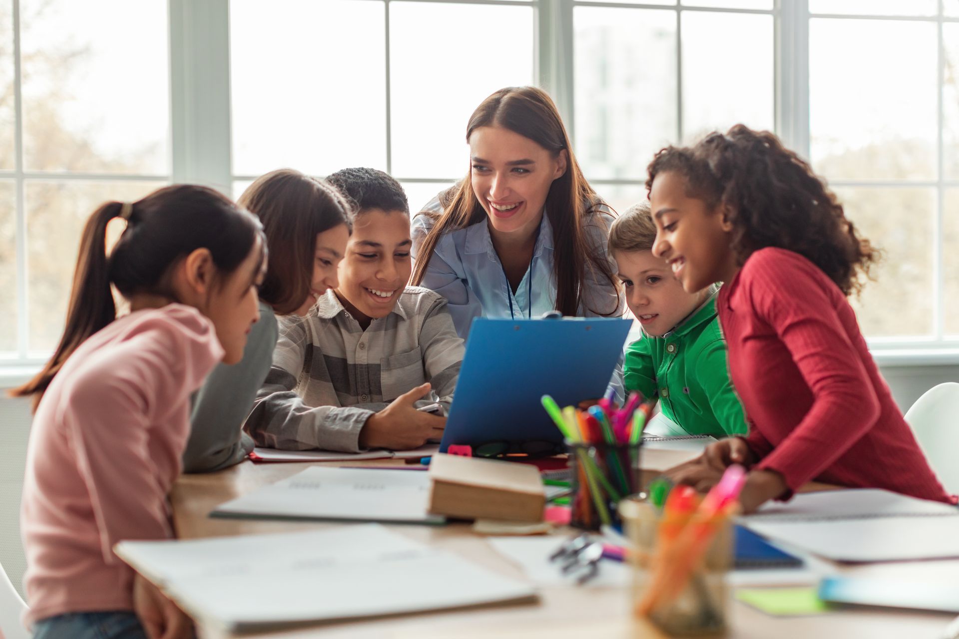 Teacher and students looking at a laptop together at a table, smiling in a bright classroom.