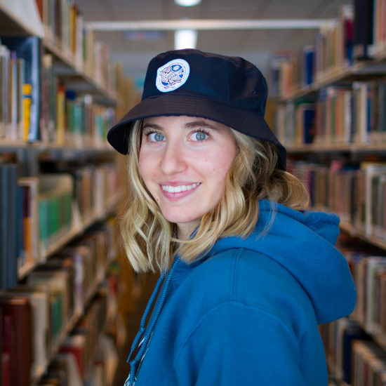 A woman wearing a hat and a blue hoodie is standing in a library.