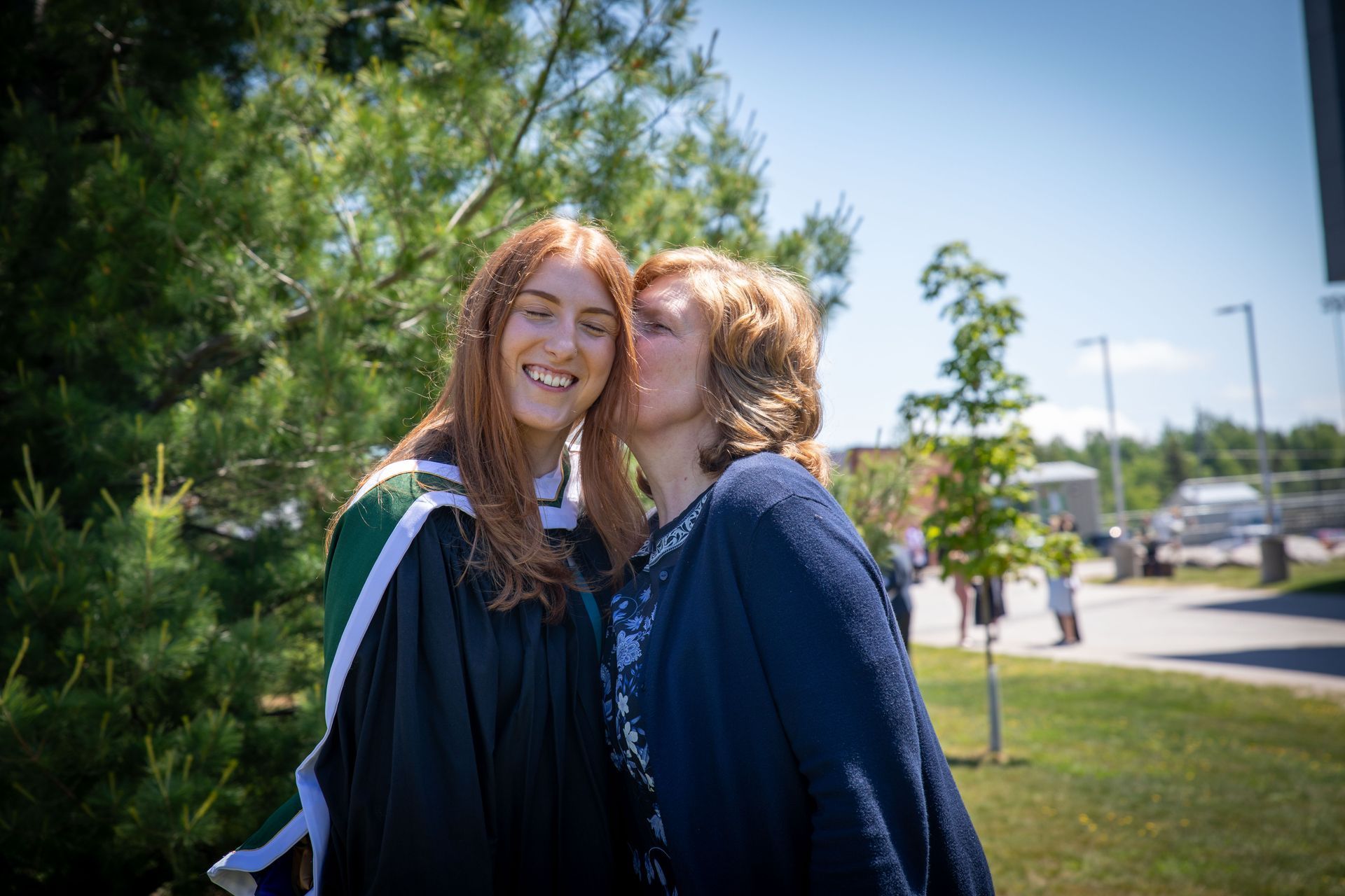 Woman in graduation gown smiles as another woman kisses her cheek outdoors.