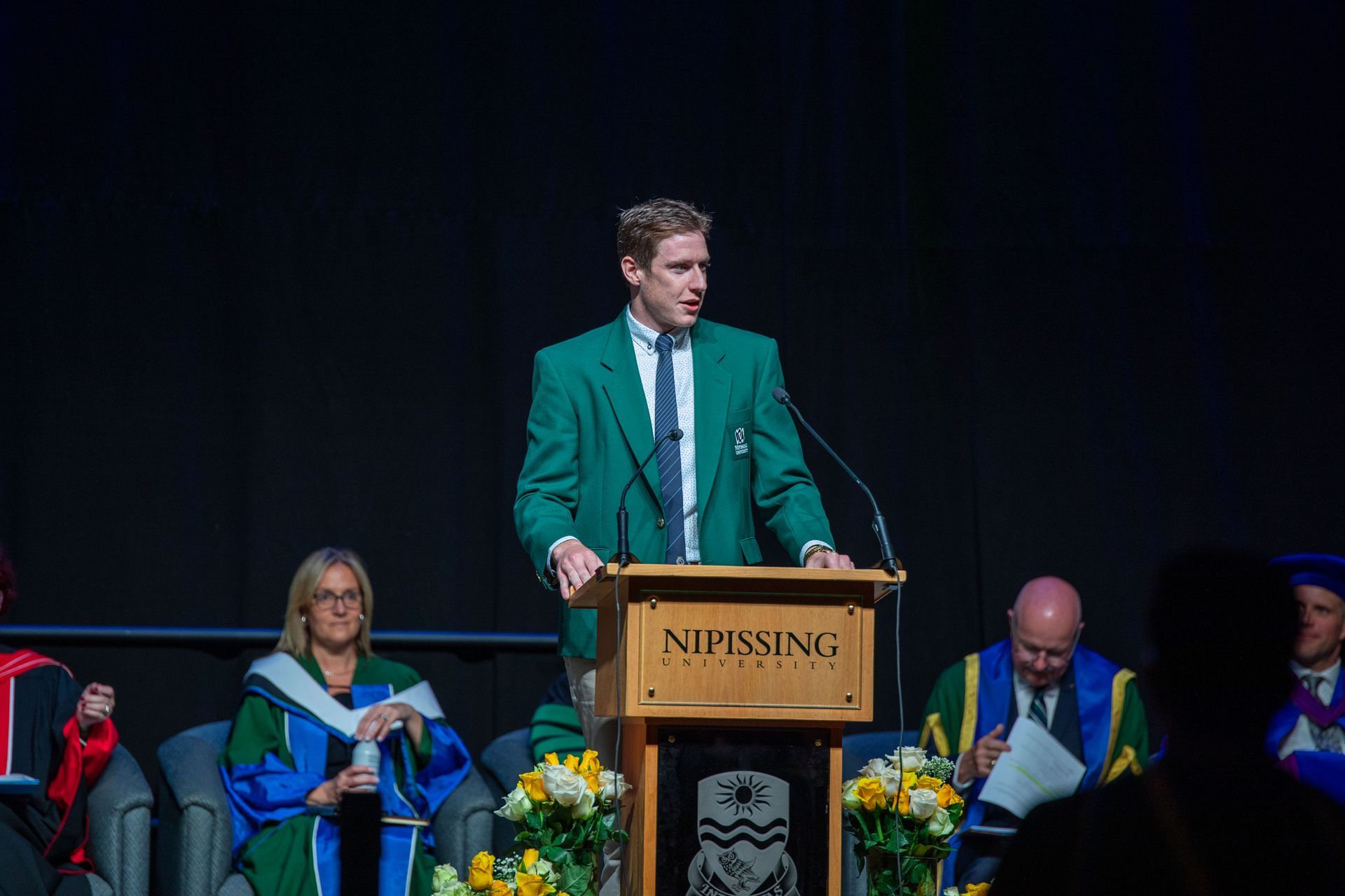 A man in a green jacket is standing at a podium giving a speech.