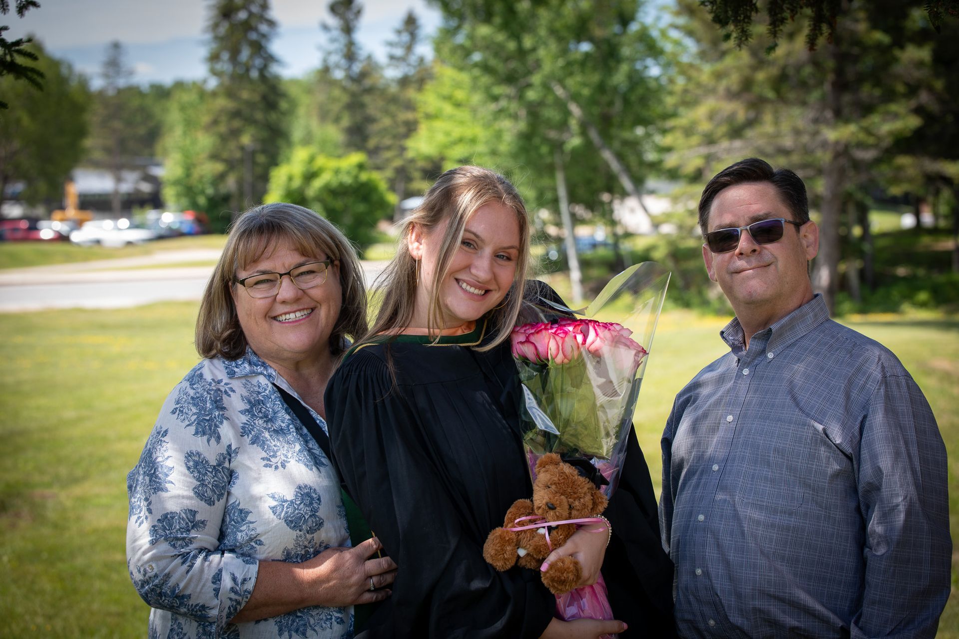 A woman is holding a bouquet of flowers and a teddy bear while posing for a picture with her parents.
