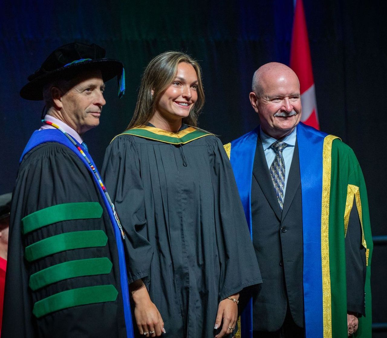 Woman in graduation gown with two men in academic robes at a ceremony; Canadian flag in background.