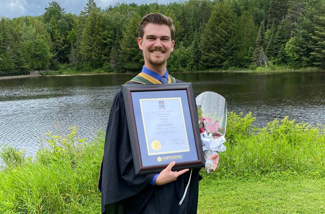 A person in a graduation gown holds a diploma and flowers near a lake.