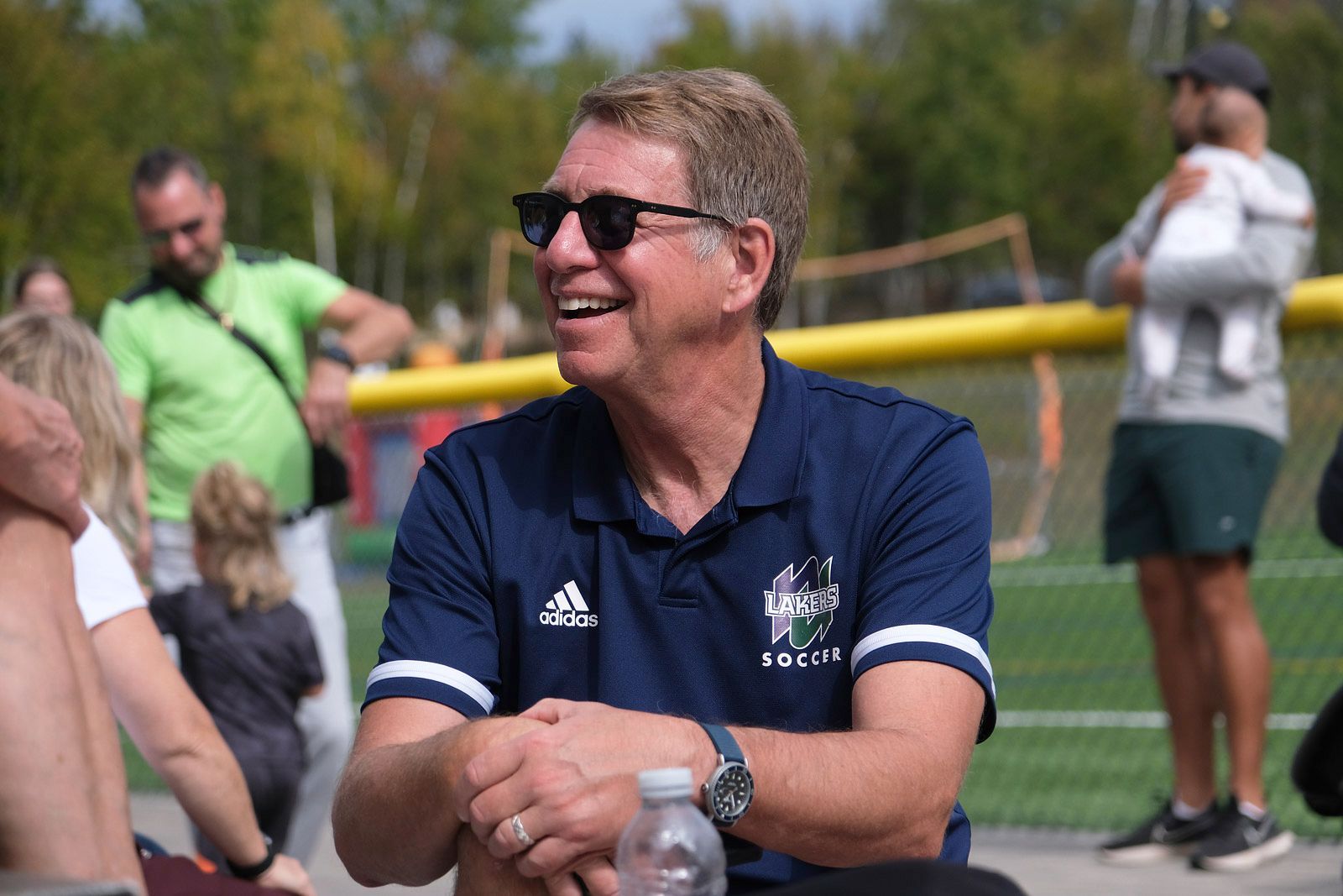 Man wearing sunglasses, smiling, in navy soccer shirt, sitting at a field.