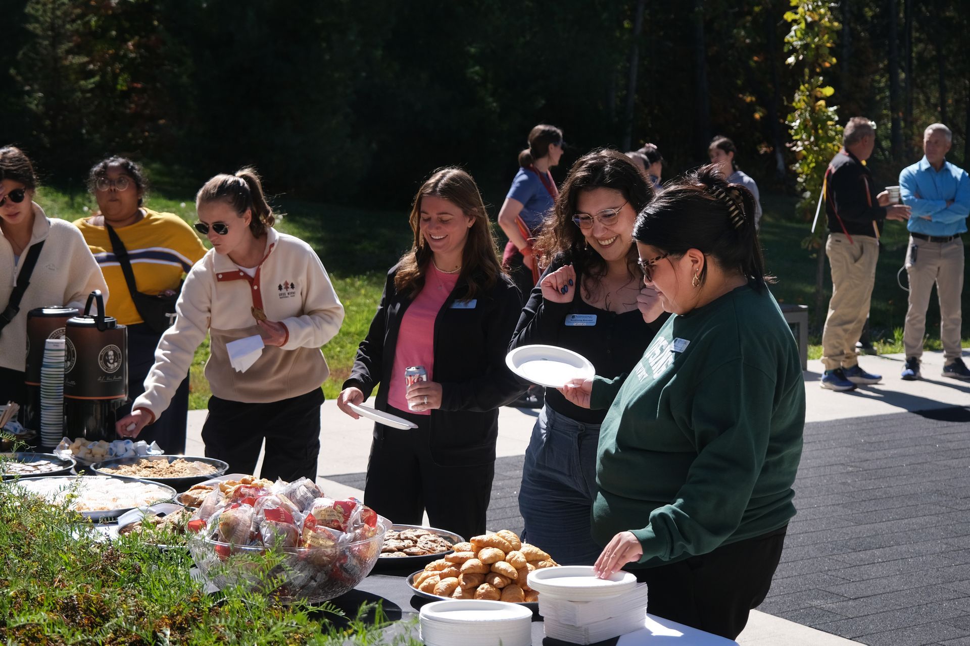 People at an outdoor food table, choosing food and socializing on a sunny day.