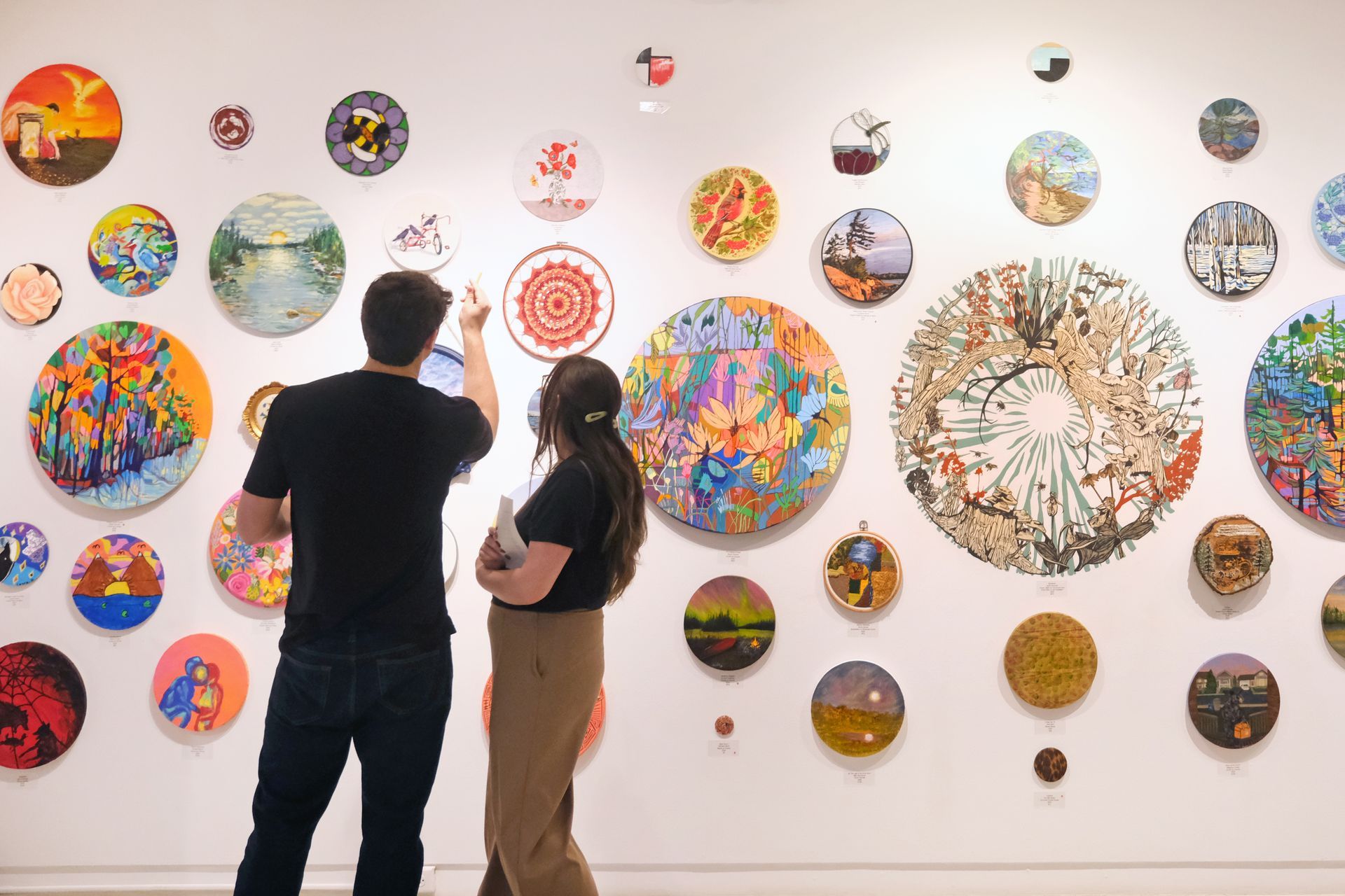 Two people in gallery viewing colorful circular artwork on white wall.