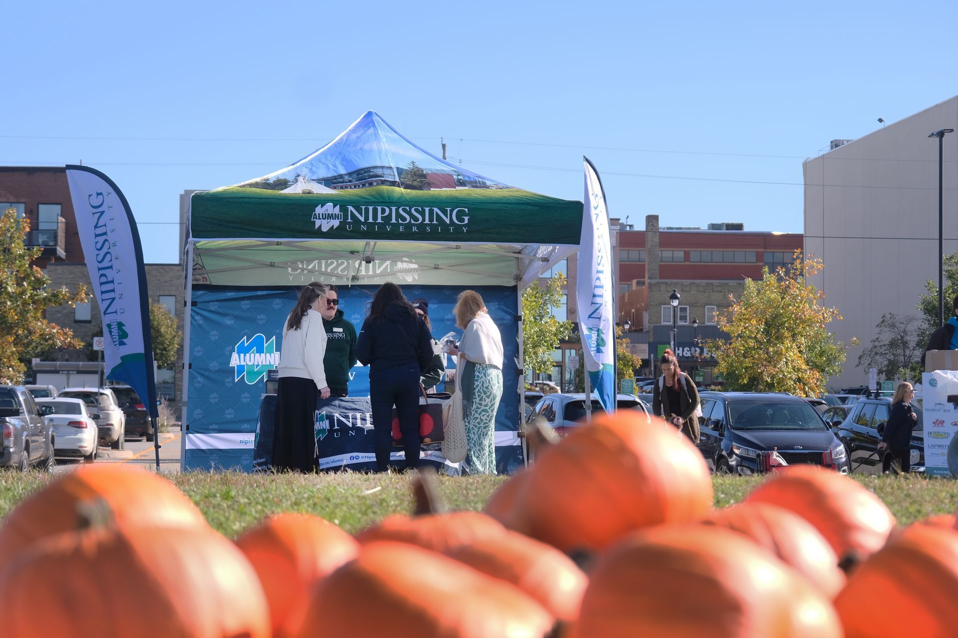 Pumpkins in foreground, Nipissing University tent with people, cars, and buildings.