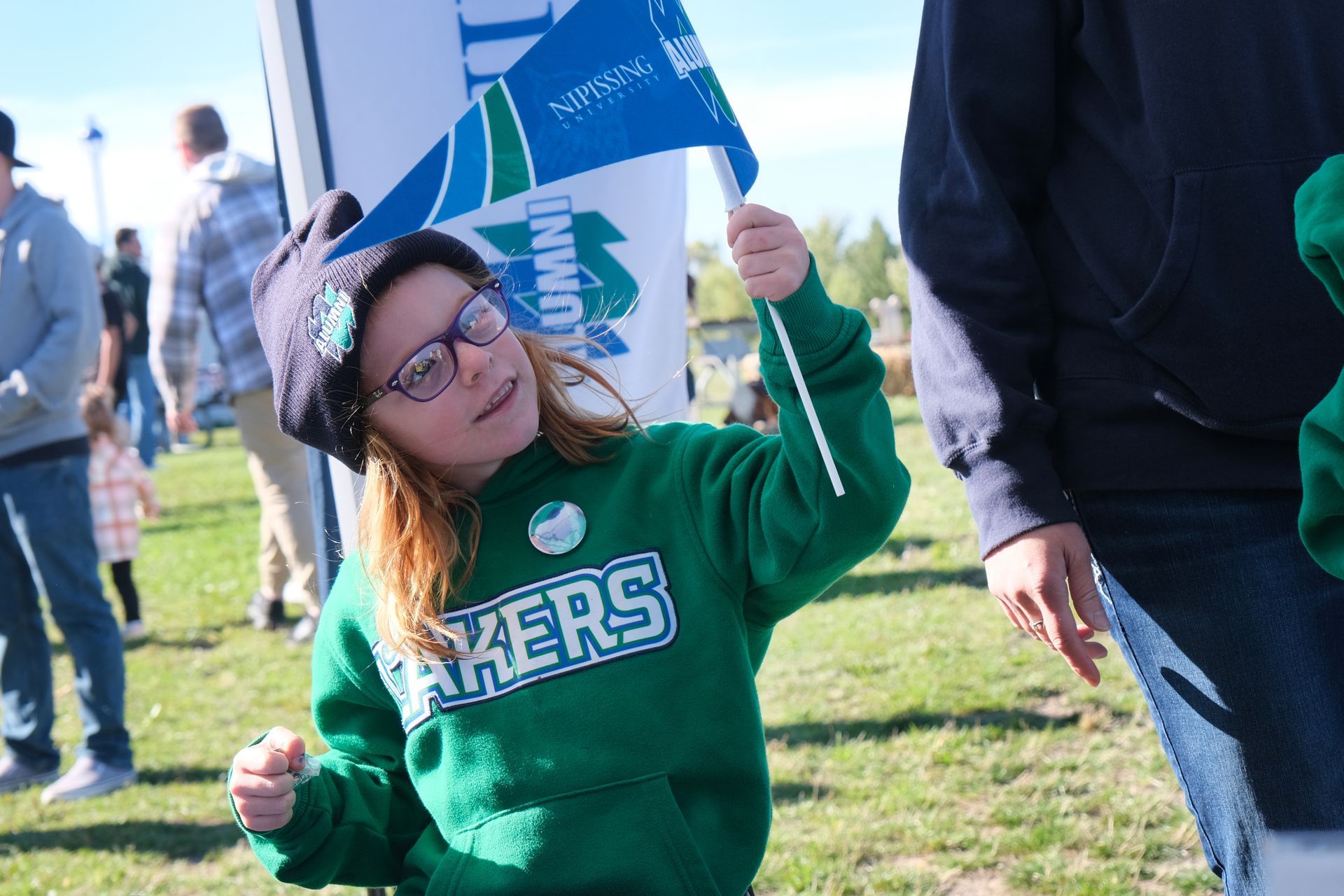 Young girl wearing glasses and a green hoodie waves a small flag at an outdoor event.