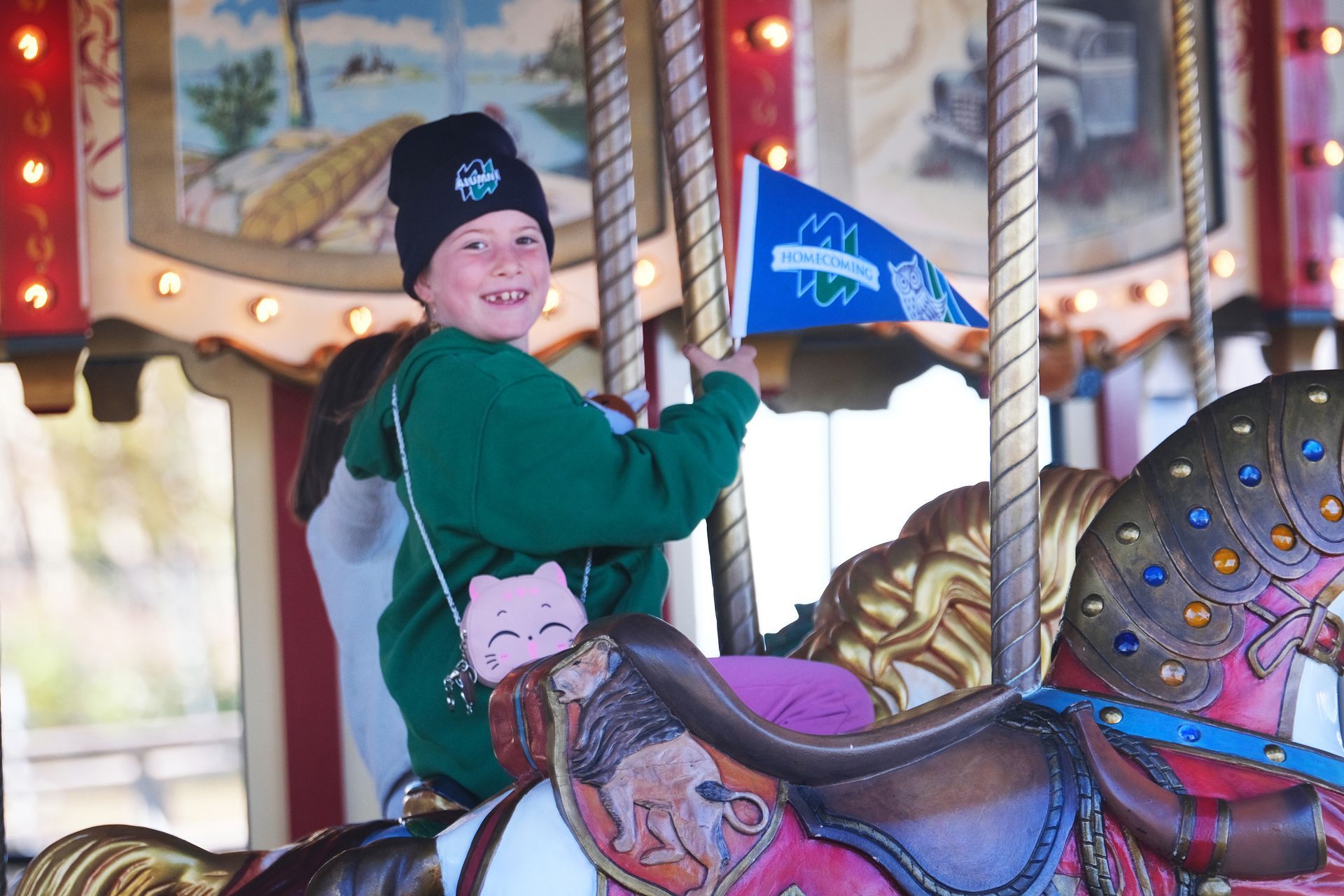 Child on a carousel horse, smiling, holding a flag. Wearing green sweatshirt, black hat, and a pink purse.