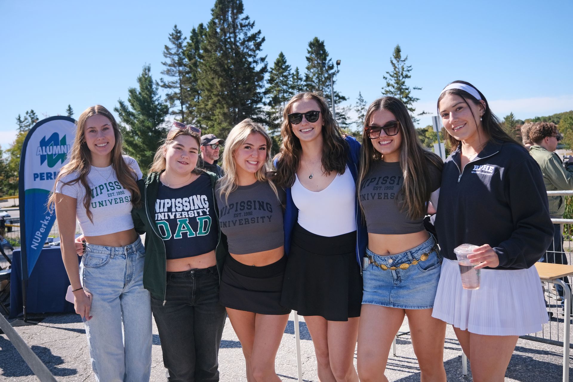 Six young women smiling, posing outdoors in casual outfits. Sunny day, trees in the background.