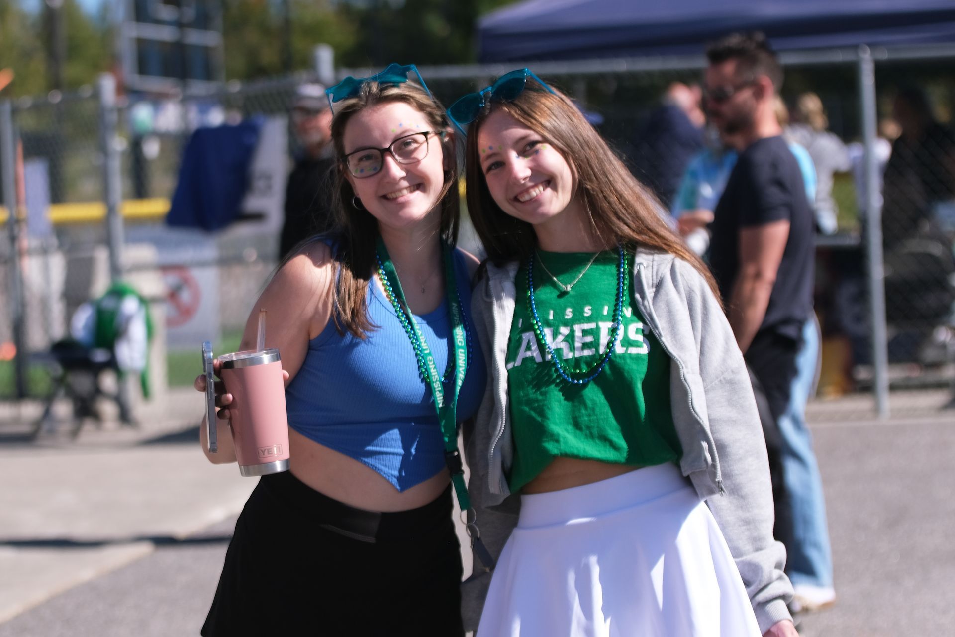 Two smiling young women in green and blue, at an outdoor event, one holding a drink.