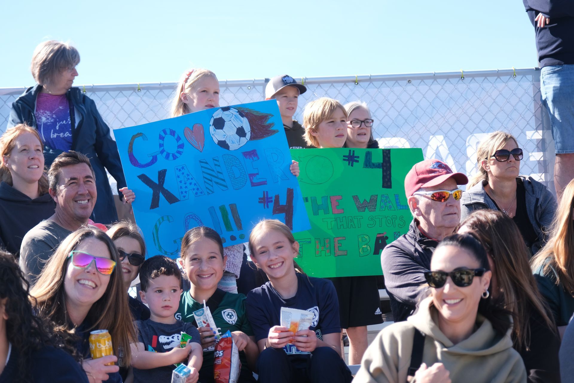 Spectators cheer at an outdoor soccer game, holding signs with soccer ball and player number 4, sunny day.