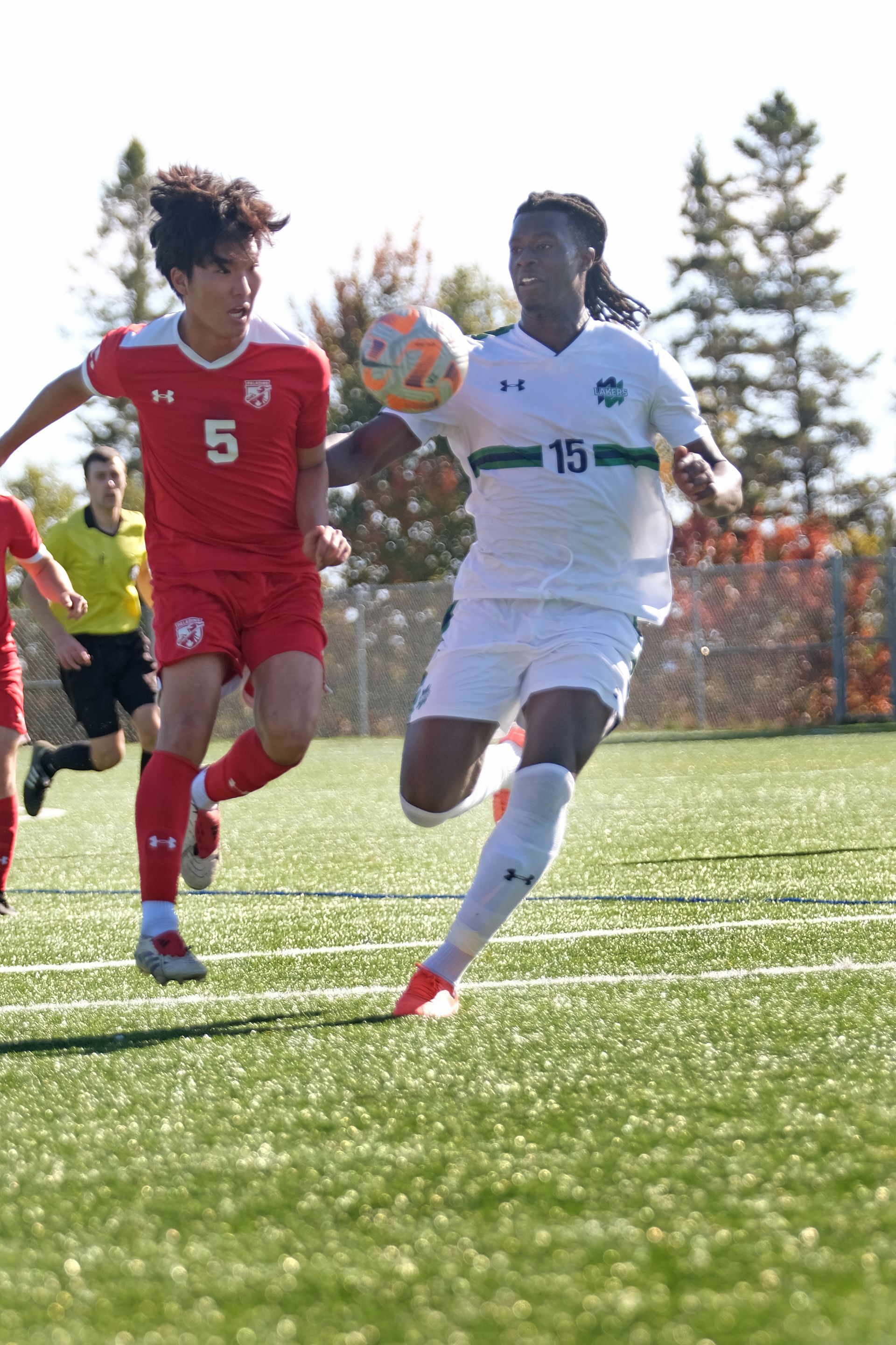 Soccer players in action: one in white, controlling the ball; one in red, running to intercept. Green field, sunny day.