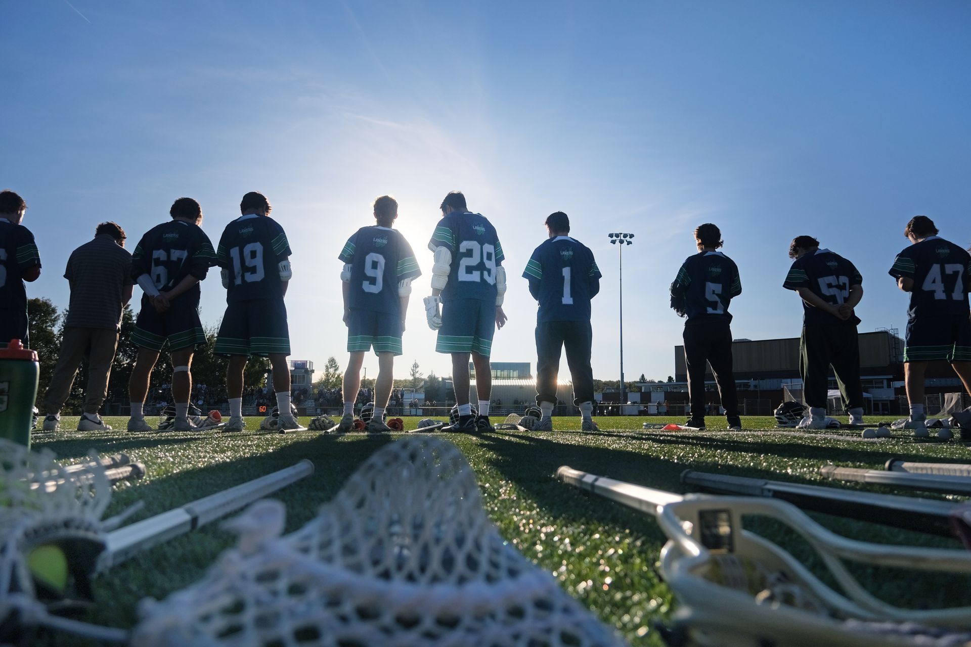 Lacrosse team stands on field, silhouetted against the sun; lacrosse sticks in foreground.