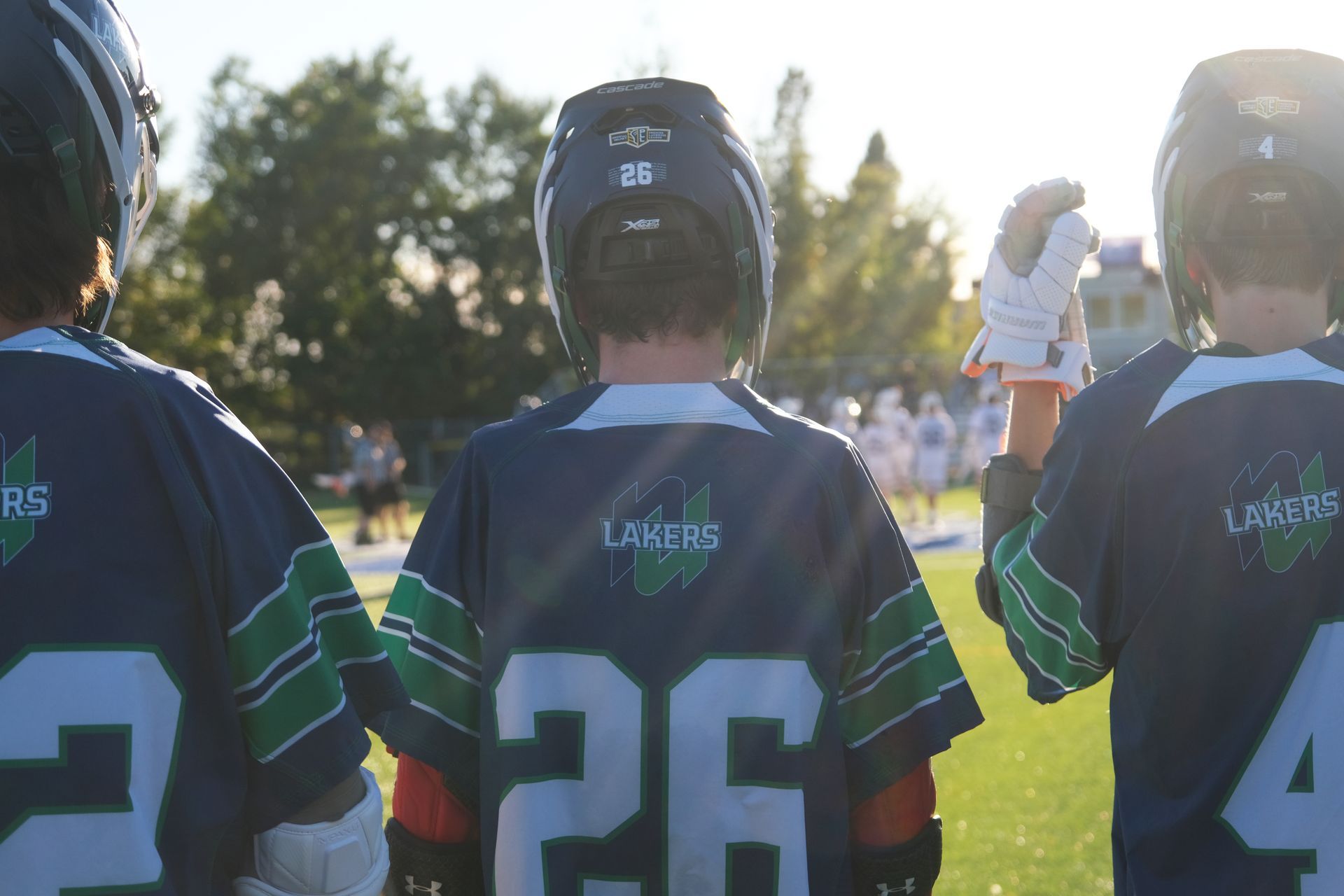 Lacrosse players in dark jerseys stand on a field. The jersey reads 