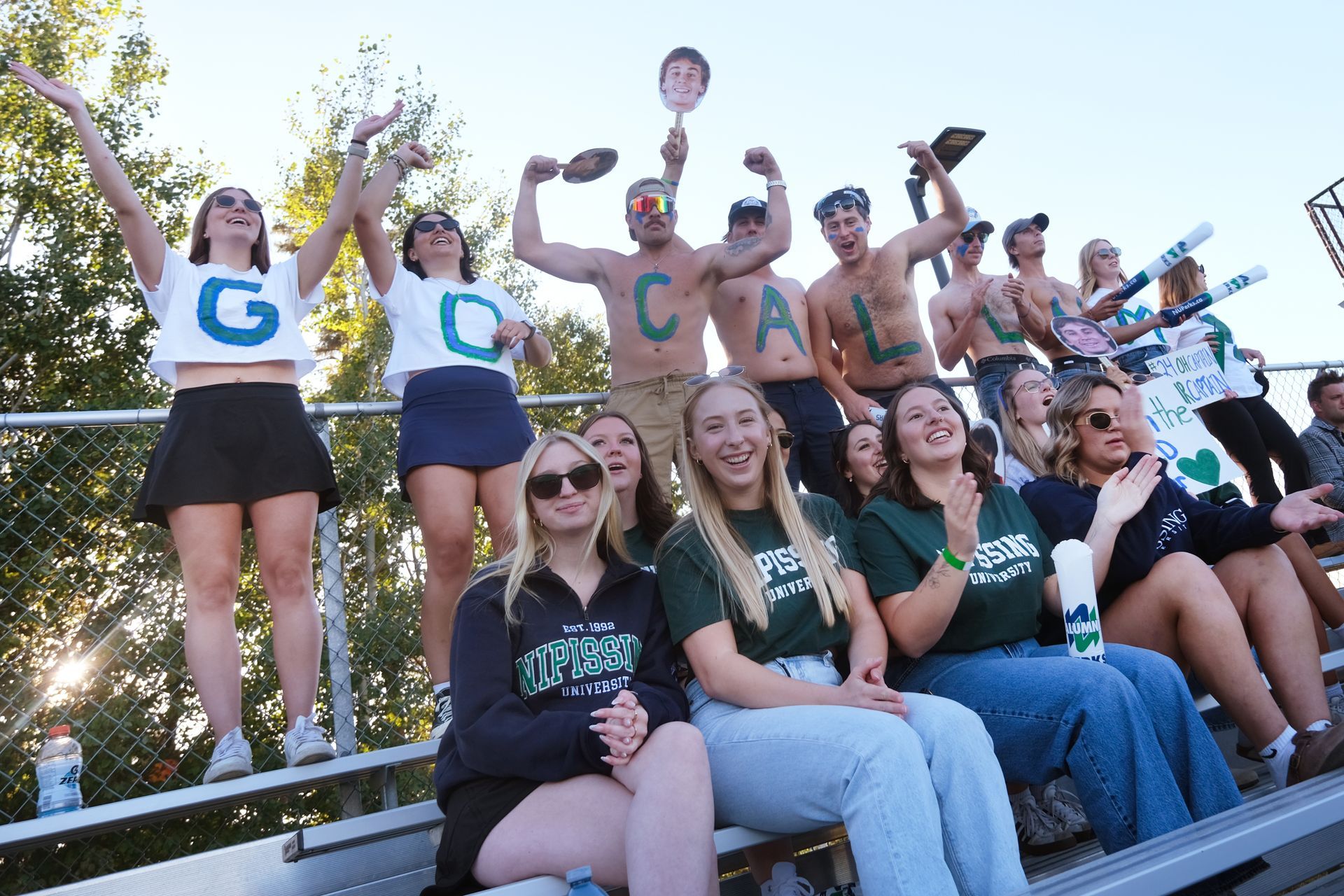 Fans cheering at a stadium, some shirtless and holding signs. 