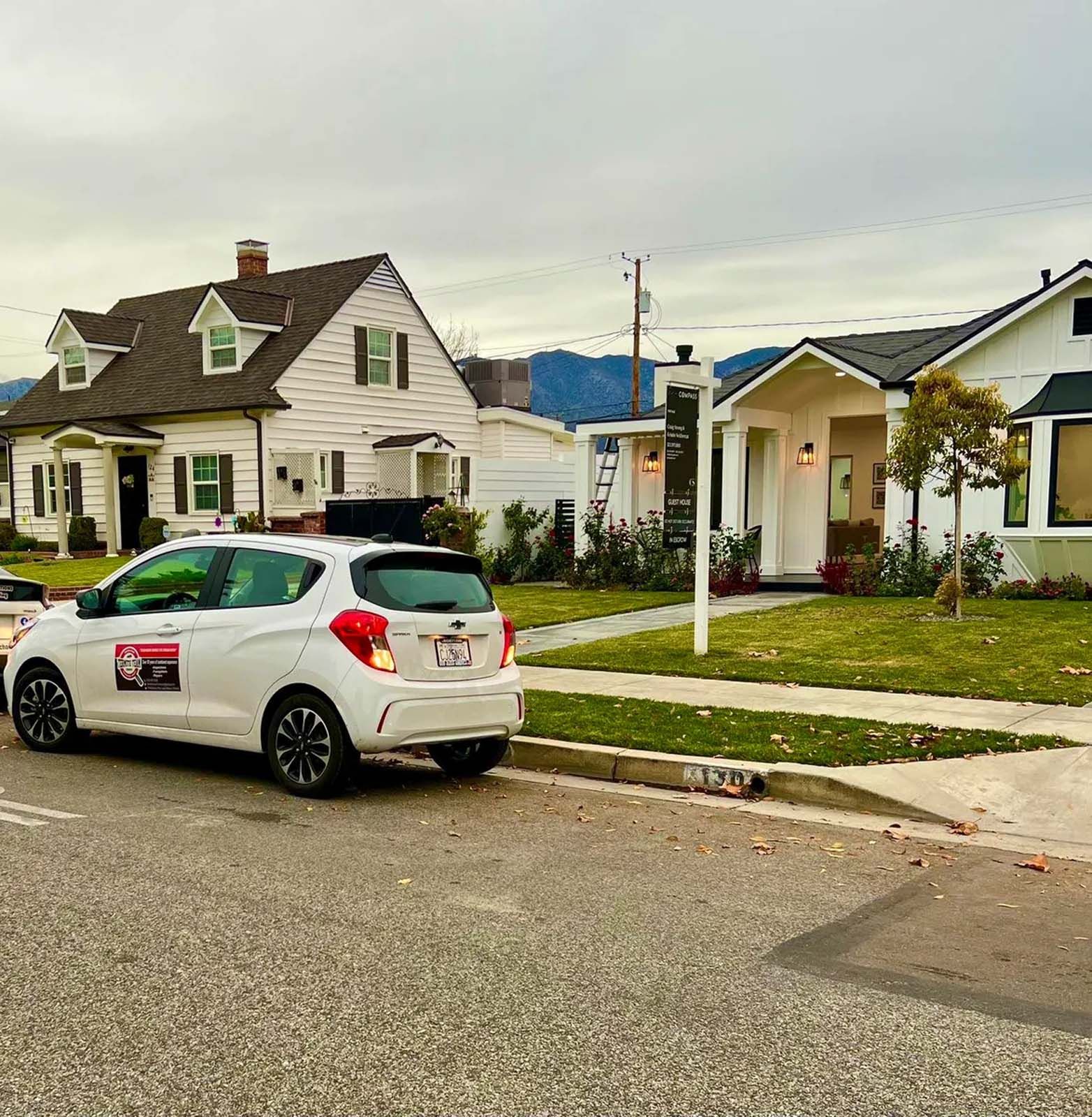 A white car is parked on the side of the road in front of a row of houses.