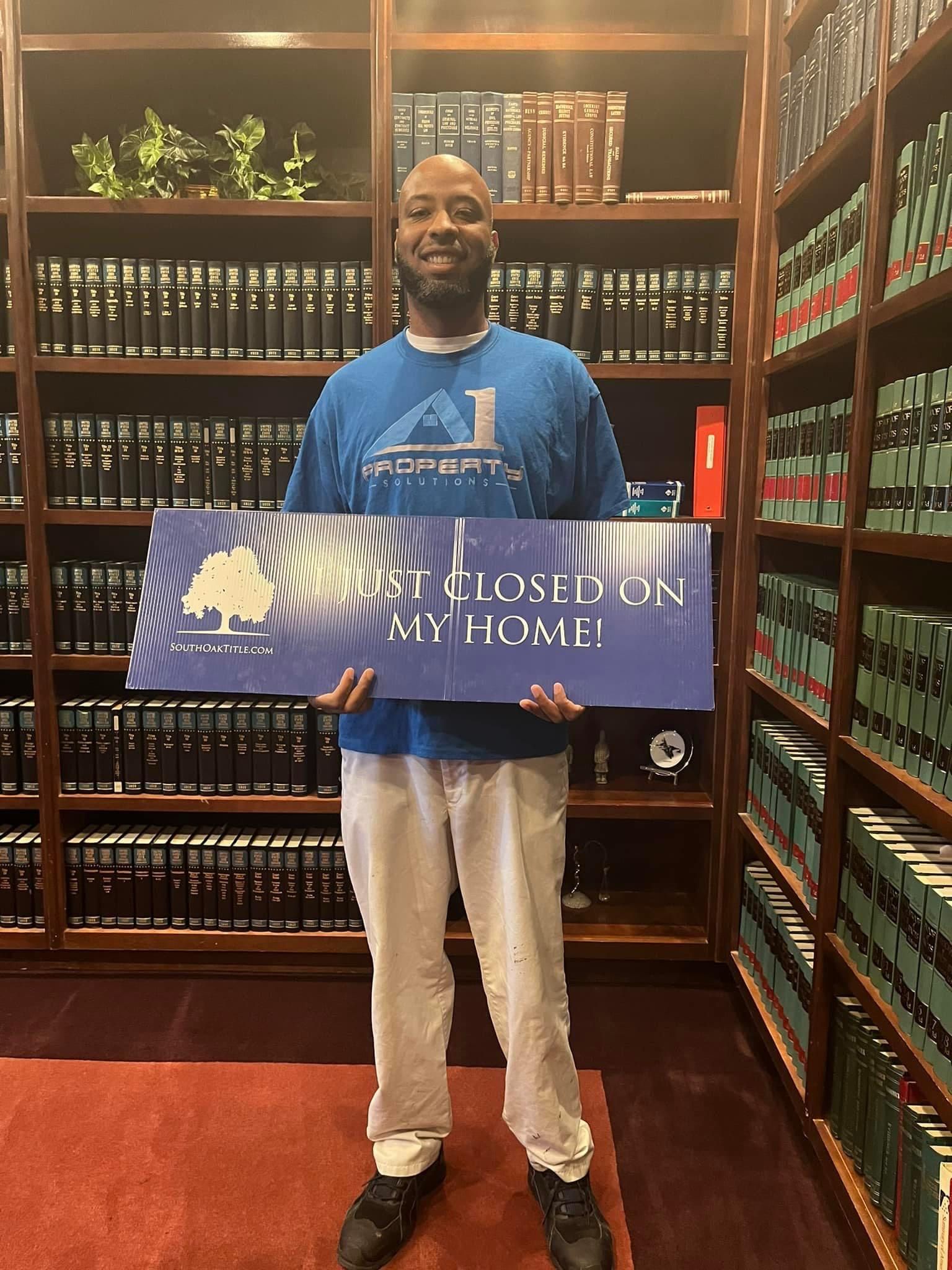 A man in a blue shirt is holding a sign in front of a bookshelf.