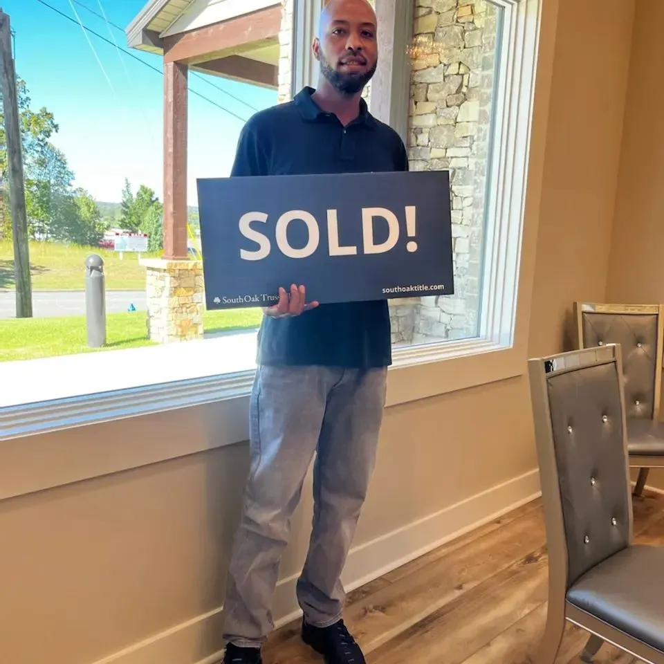 A man is holding a sold sign in front of a window.