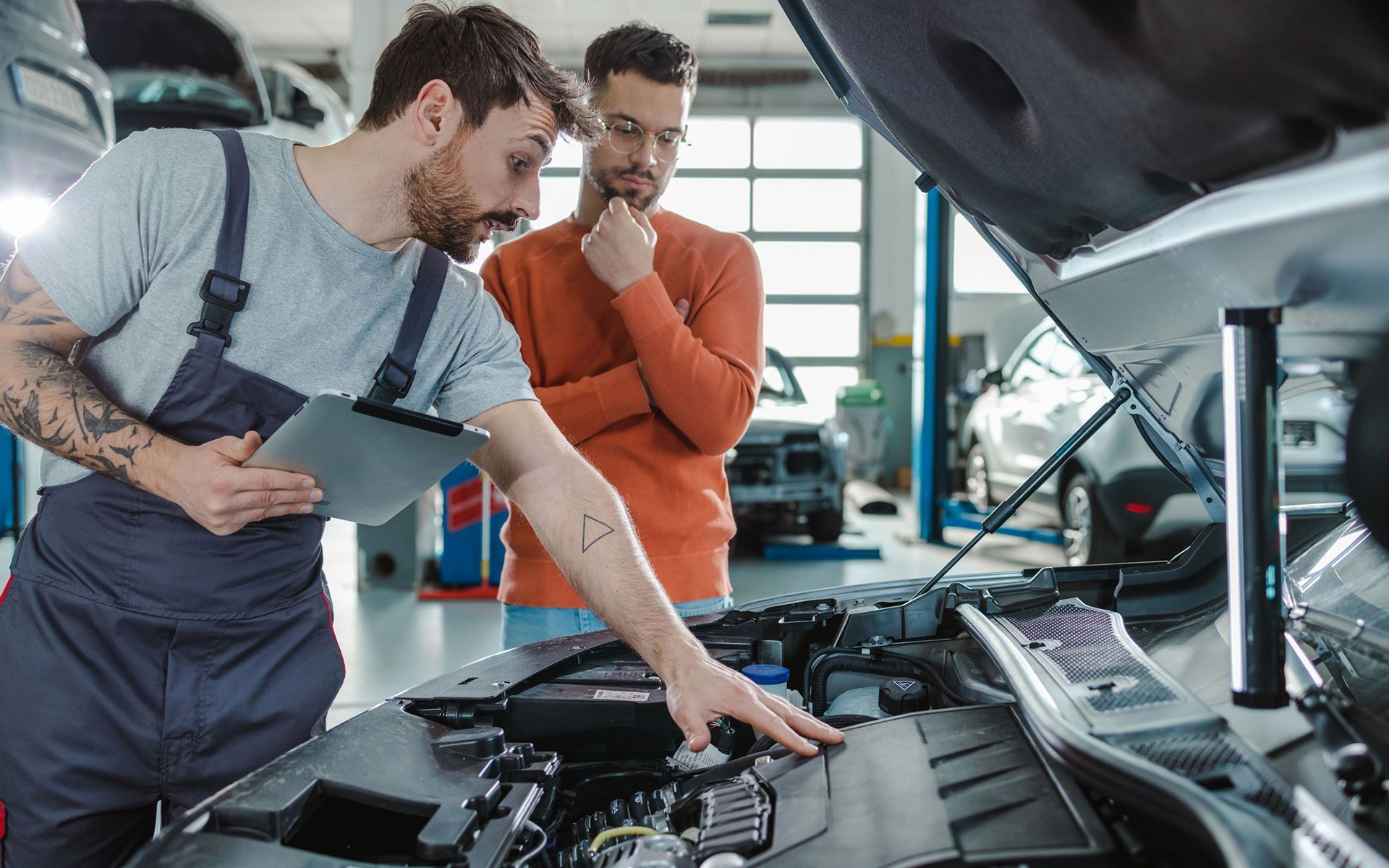 Two men are looking under the hood of a car in a garage.