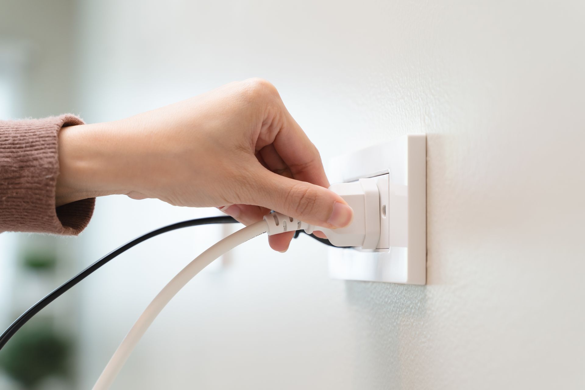 Hand plugging a white power cord into an electrical outlet on a white wall.