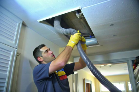 Worker wearing yellow gloves cleans a ceiling vent with a hose in an indoor hallway.