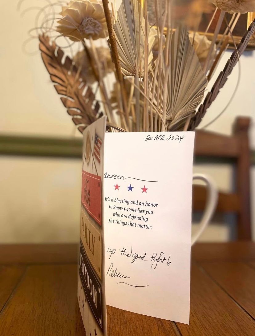 Greeting card in vase with dried flowers, on a wood table. Card has text, and red, white, and blue colors.