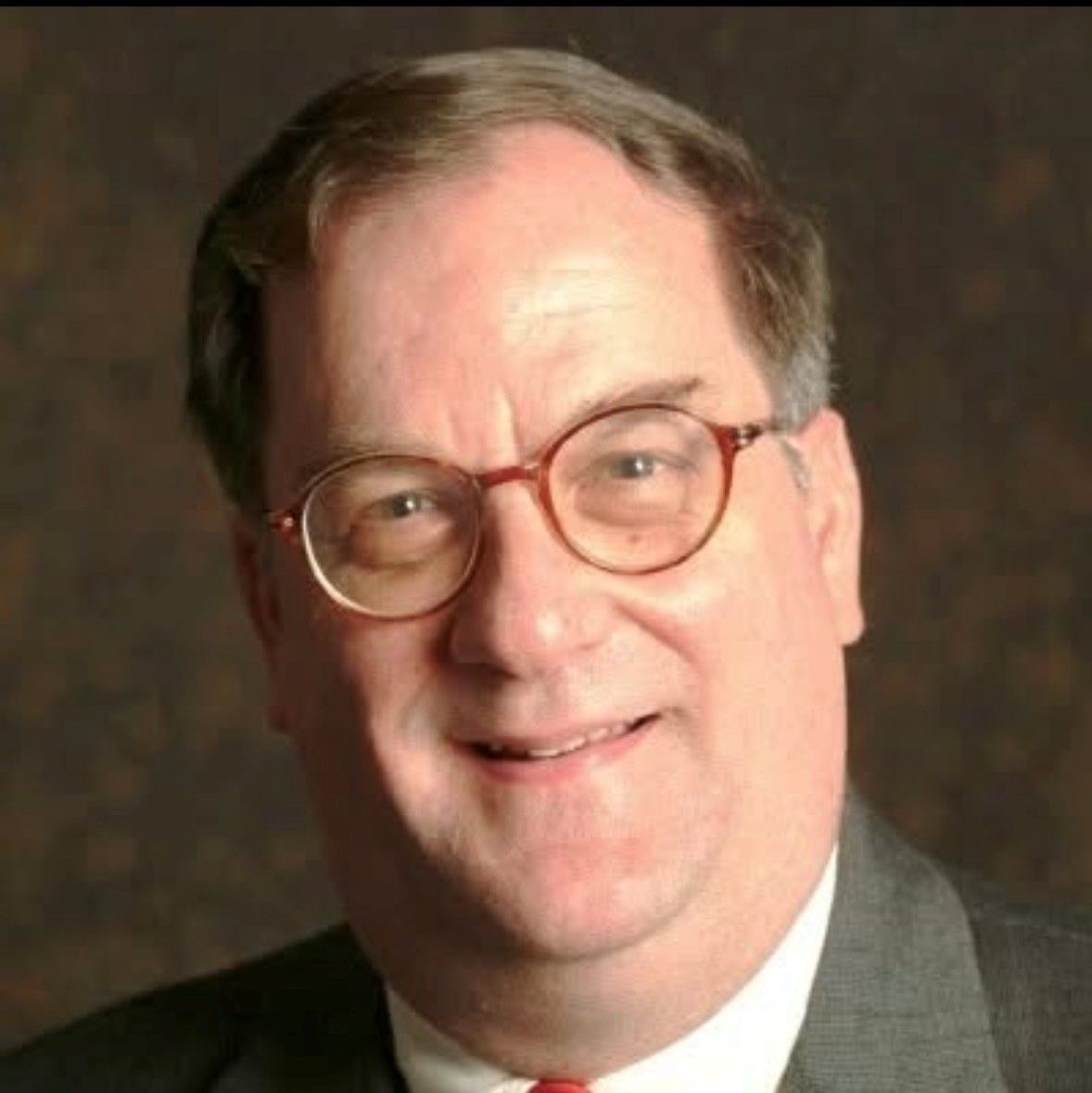 A professional headshot of a person wearing glasses and a gray suit jacket with a white shirt and red tie.